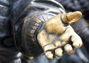 A close-up of the left hand of a bronze statue. The hand is a polished golden colour, in contrast to the rest of the statue which is a dark brown colour.